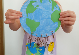 Dziecko trzyma w dłoniach wyciętą z papieru kulę ziemską. Do kuli przyczepione są na sznureczkach papierowe przywieszki: niebieska kropla, zielone drzewko, jasnoniebieska butelka oraz kolorowe trapezy symbolizujące kontenery na śmieci.